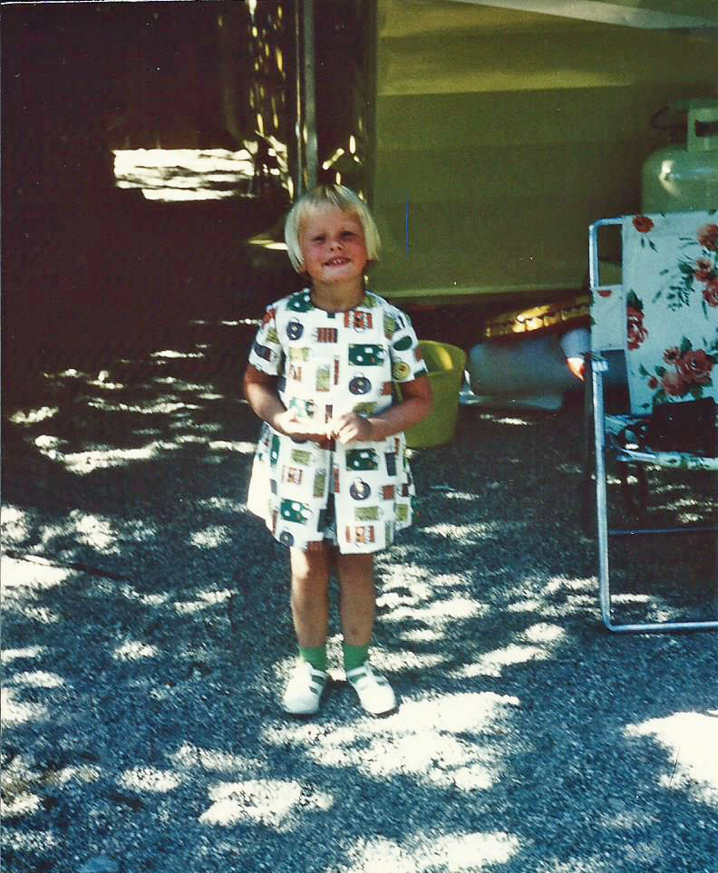 Three year old girl standing in front of a travel trailer and wearing a white printed dress made for her by her mother. 